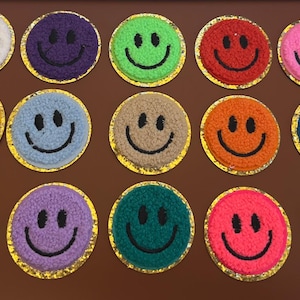 May include: A collection of colorful smiley face patches. Each patch features a different color smiley face with a black stitched mouth and eyes, surrounded by a gold border. The patches are arranged on a brown surface.