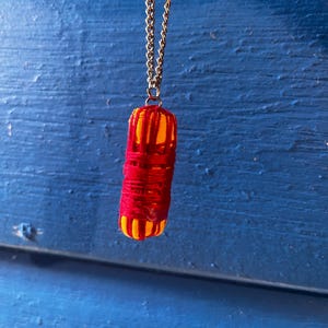 May include: A pendant necklace with a cylindrical charm wrapped in red thread. The charm has yellow and orange accents and hangs from a silver-coloured chain. The background is a textured blue surface.