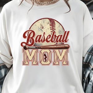 May include: White t-shirt featuring a baseball design. The design includes a baseball, a bat, and the words "Baseball MOM" in red and tan lettering. The baseball has red stitching.