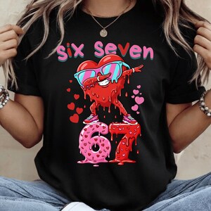 Può includere: T-shirt nera con una grafica a forma di cuore rosso con occhiali da sole che fa dab sui numeri 6 e 7. Il testo "Six Seven" è sopra il cuore, con piccoli cuori rosa sparsi.
