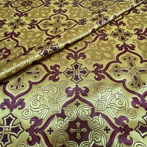 May include: Gold-colored fabric with a repeating pattern of crosses and fleur-de-lis in a deep burgundy color. The fabric has a textured, embossed appearance, with folds adding depth to the design. The overall aesthetic suggests a religious or ceremonial theme.