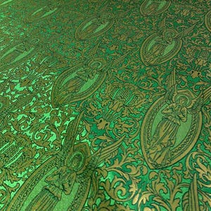 May include: Green fabric with a repeating pattern of angels in oval frames and ornate gold floral designs. The fabric has a textured appearance and a slight sheen, creating a luxurious look. The overall design is detailed and intricate.