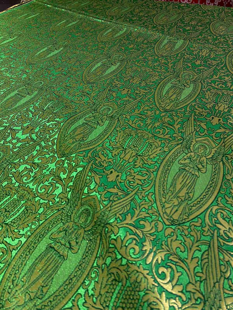 May include: Green fabric with a repeating pattern of angels in oval frames and ornate gold floral designs. The fabric has a textured appearance and a slight sheen, creating a luxurious look. The overall design is detailed and intricate.