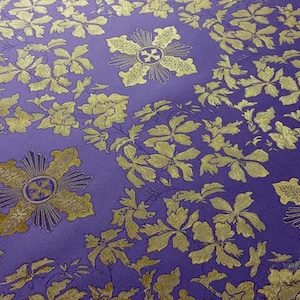 May include: A close-up of a purple fabric with a repeating pattern of gold floral and cross-like designs. The gold elements have a metallic sheen, contrasting with the matte purple background. The pattern is dense and detailed.