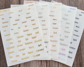 May include: A set of eight sheets of clear sticker paper with gold foil lettering. Each sheet features a different month of the year, with the months repeated multiple times on each sheet. The sheets are arranged in a fan shape, with the top sheet showing the month of January.