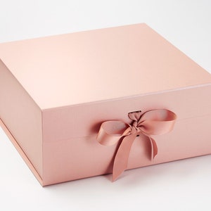 May include: A pale pink gift box with a matching satin ribbon tied in a bow. The box is rectangular with a magnetic closure and a matte finish. The box is empty.