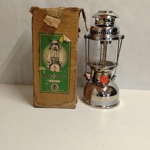 Könnte beinhalten: Eine Vintage-Laterne aus Chrom und Glas mit einer transparenten Glaskugel und einem roten Ventil. Die Laterne steht neben der originalen Pappschachtel, die ein grünes und braunes Etikett mit dem Text "HIPOLITO AUTOMATIC" hat.