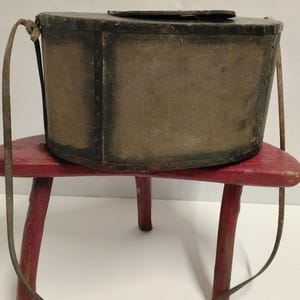 May include: An antique rectangular box with a hinged lid and a shoulder strap. The box has a dark frame and a mesh-like fabric body. It sits on a small, red, three-legged stool. The strap is a light brown colour.