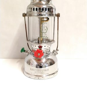 Könnte beinhalten: Vintage silberfarbene Laterne mit rotem Deckel und einem Zylinder aus klarem Glas. Die Laterne hat einen grünen und einen roten Knopf sowie einen Griff. Die Worte "LIGHT BRAND" sind auf der Basis sichtbar.