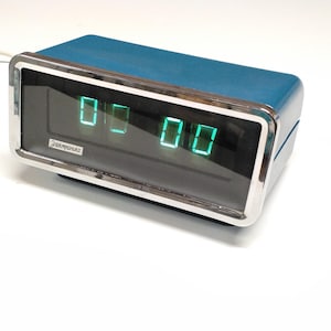 Pode incluir: Um relógio digital vintage azul-petróleo com uma moldura prateada. O relógio mostra a hora "00:00" em algarismos verdes. A marca "Электроника" é visível na frente. O relógio é retangular e tem um cabo de alimentação branco.