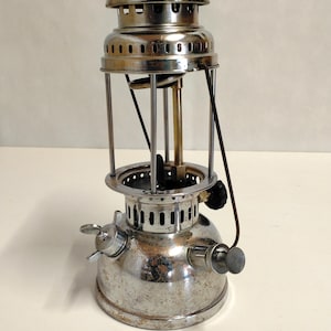 Könnte beinhalten: Vintage silberfarbene Laterne mit rundem Sockel und zylindrischem Oberteil. Die Laterne verfügt über einen Griff, ein Kraftstoffventil und ein Glasgehäuse. Das Metall hat ein verwittertes Aussehen, was auf Alter und Nutzung hindeutet. Für Außen- oder Notbeleuchtung.