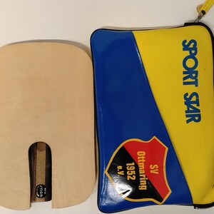 May include: A light-colored wooden ping pong paddle with a U-shaped handle is next to a blue and yellow zippered case. The case has the words "SPORT STAR" printed on it, along with a logo that says "SV Ottmaring 1952 e.V."