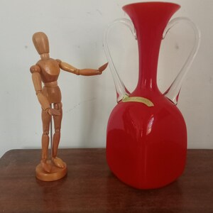 Op de afbeelding: Een rode glazen vaas met twee transparante glazen handvatten. De vaas heeft een ronde bodem en een smalle hals.