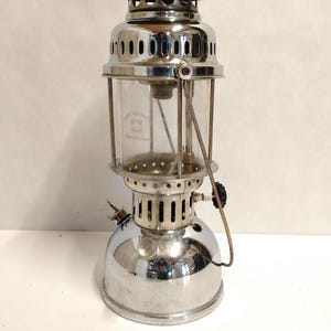 May include: A vintage chrome lantern with a clear glass globe. The lantern has a rounded top and base, with ventilation holes. The glass globe has text that reads "Apex & Denmark". A handle is attached. Measures approximately 30 cm in height.