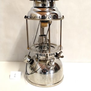 Könnte beinhalten: Eine silberfarbene Vintage-Laterne mit einem transparenten Glaszylinder. Die Laterne hat einen oberen Griff, eine Tankanzeige und den Text "HPS G" auf der Basis. Diese klassische Laterne ist für den Außenbereich konzipiert.