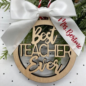 May include: A wooden ornament with the words "Best Teacher Ever" in red lettering. The ornament is decorated with a white satin bow and a white ribbon with the text "Mrs. Beestone" in red lettering.
