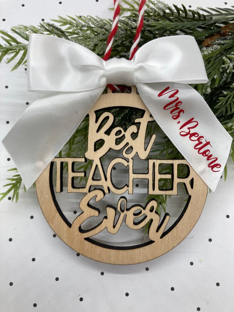 May include: A wooden ornament with the words "Best Teacher Ever" in red lettering. The ornament is decorated with a white satin bow and a white ribbon with the text "Mrs. Beestone" in red lettering.