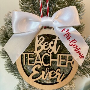May include: A wooden Christmas ornament with the words "Best Teacher Ever" cut out. The ornament is decorated with a white satin bow and a red and white ribbon. The ribbon has the name "Mrs. Bertone" written on it.
