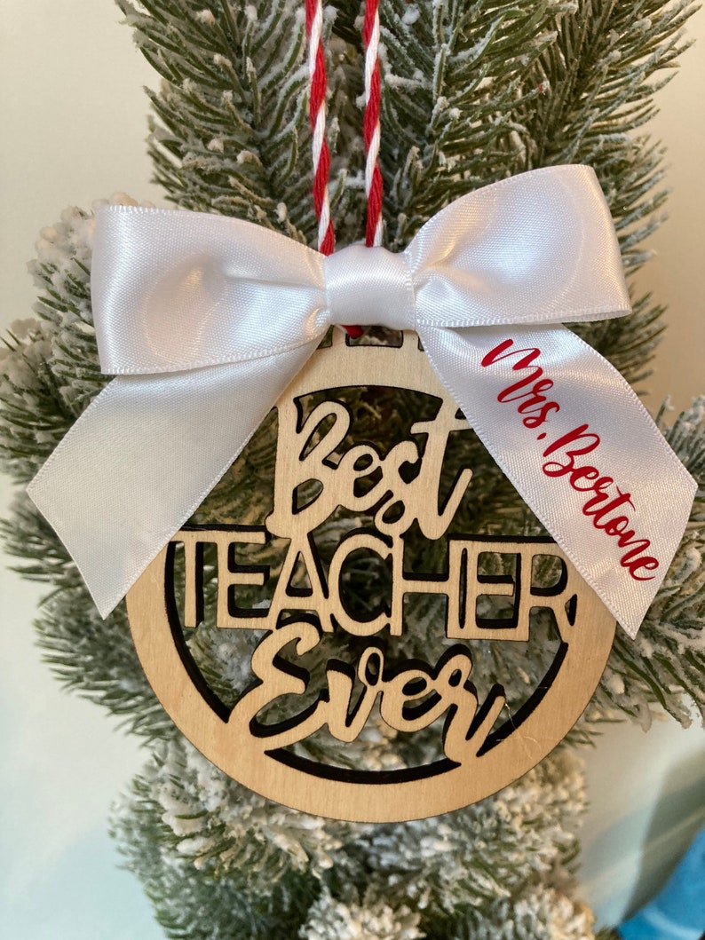 May include: A wooden Christmas ornament with the words "Best Teacher Ever" cut out. The ornament is decorated with a white satin bow and a red and white ribbon. The ribbon has the name "Mrs. Bertone" written on it.