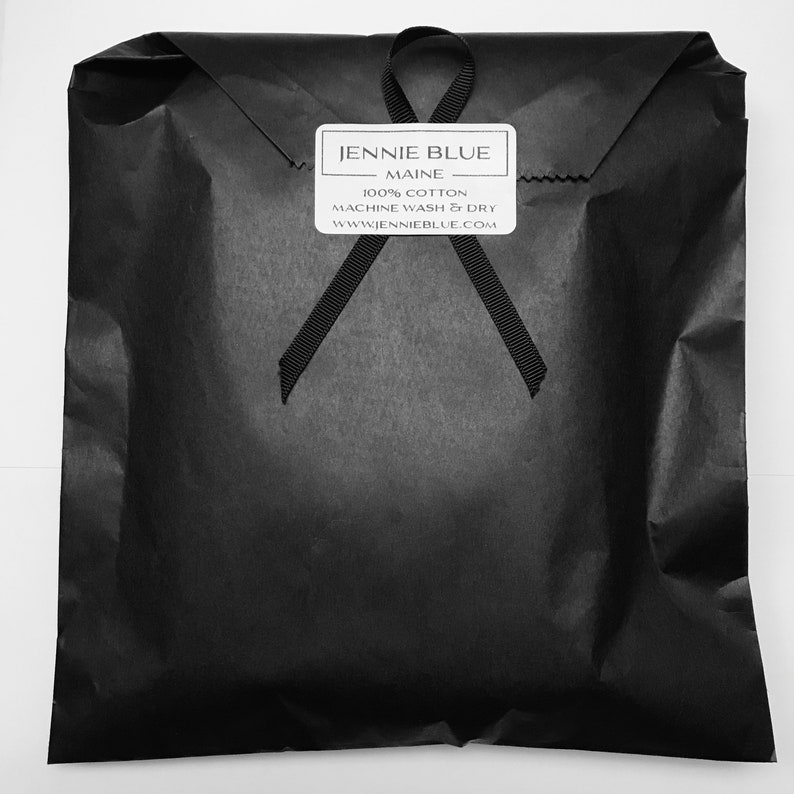 Pode incluir: Um saco de papel preto com uma fita preta amarrada em volta. O saco tem um r&oacute;tulo branco com o texto "JENNIE BLUE - MAINE - 100% COTTON MACHINE WASH & DRY WWW.JENNIENBLUE.COM"