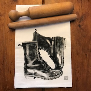May include: A white tea towel with a black and white illustration of two black leather boots with laces. The boots are on a white background.