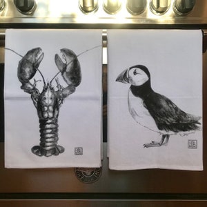 Pode incluir: Duas toalhas de cozinha brancas com ilustra&ccedil;&otilde;es em preto e branco. Uma toalha apresenta uma lagosta, a outra um papagaio-do-mar.