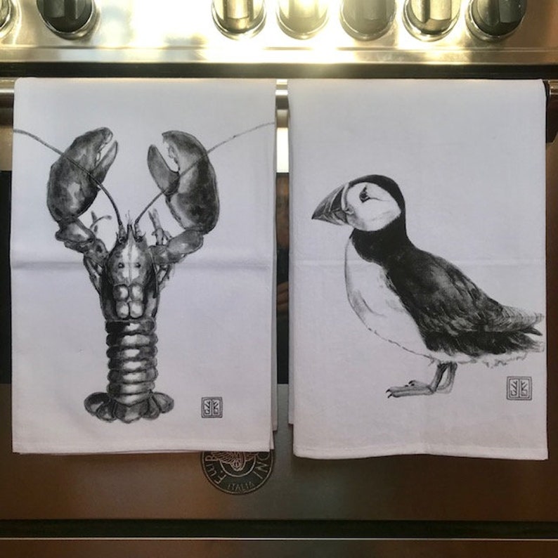 Pode incluir: Duas toalhas de cozinha brancas com ilustra&ccedil;&otilde;es em preto e branco. Uma toalha apresenta uma lagosta, a outra um papagaio-do-mar.