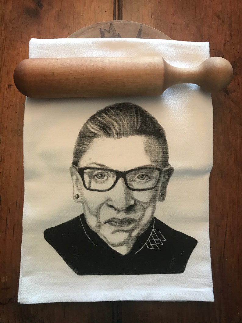 May include: A white kitchen towel with a black and white image of Ruth Bader Ginsburg. The towel is folded and lying on a wooden surface.