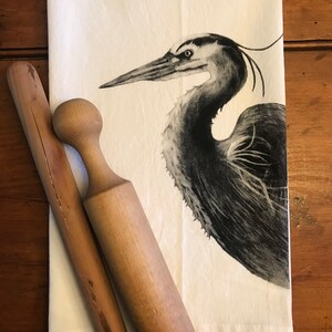 May include: A white kitchen towel with a black and white illustration of a heron. The towel is folded and lying on a wooden surface with two wooden rolling pins.