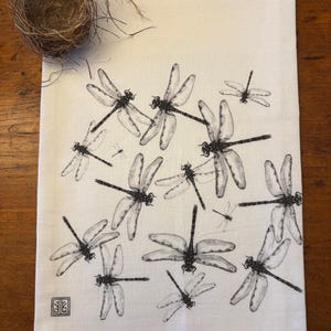 May include: White kitchen towel with a black and white dragonfly print. The towel has a small black square with a symbol in the bottom left corner.