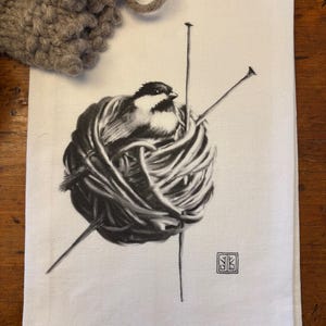 May include: A black and white illustration of a small bird sitting in a ball of yarn with two knitting needles sticking out. The illustration is on a white tea towel with a small black square with a design in the bottom right corner.