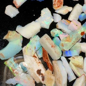 May include: A collection of raw, unpolished opal stones in various shapes and sizes. The stones feature a milky white base color with iridescent flashes of blue, green, and orange light. Some pieces show earthy brown inclusions and rough textures.