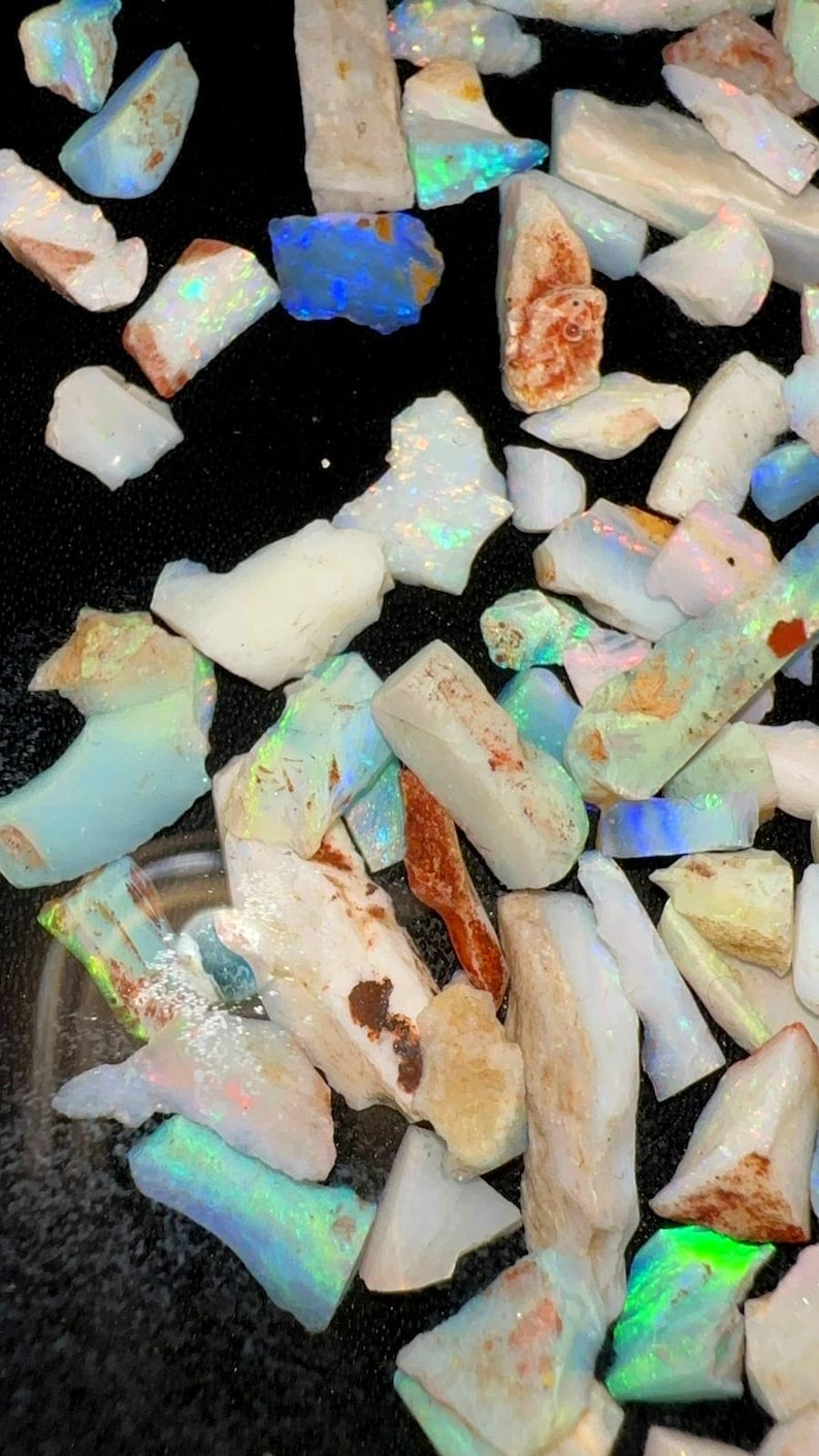 May include: A collection of raw, unpolished opal stones in various shapes and sizes. The stones feature a milky white base color with iridescent flashes of blue, green, and orange light. Some pieces show earthy brown inclusions and rough textures.