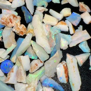 May include: A collection of raw, unpolished opal stones featuring irregular shapes and varying sizes. The stones display a milky white base color with iridescent flashes of blue, green, and orange light reflecting from their surfaces.