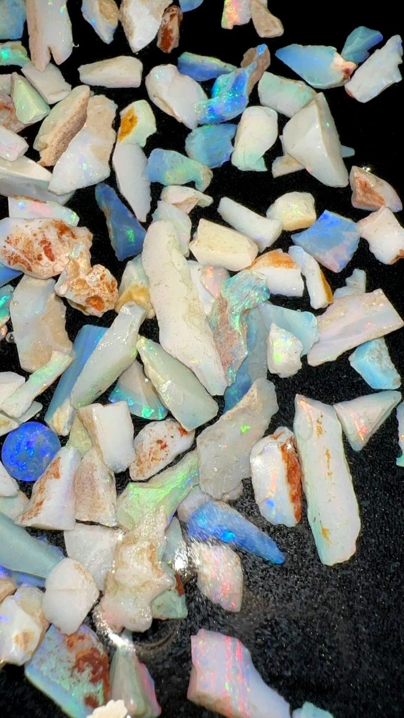 May include: A collection of raw, unpolished opal stones featuring irregular shapes and varying sizes. The stones display a milky white base color with iridescent flashes of blue, green, and orange light reflecting from their surfaces.