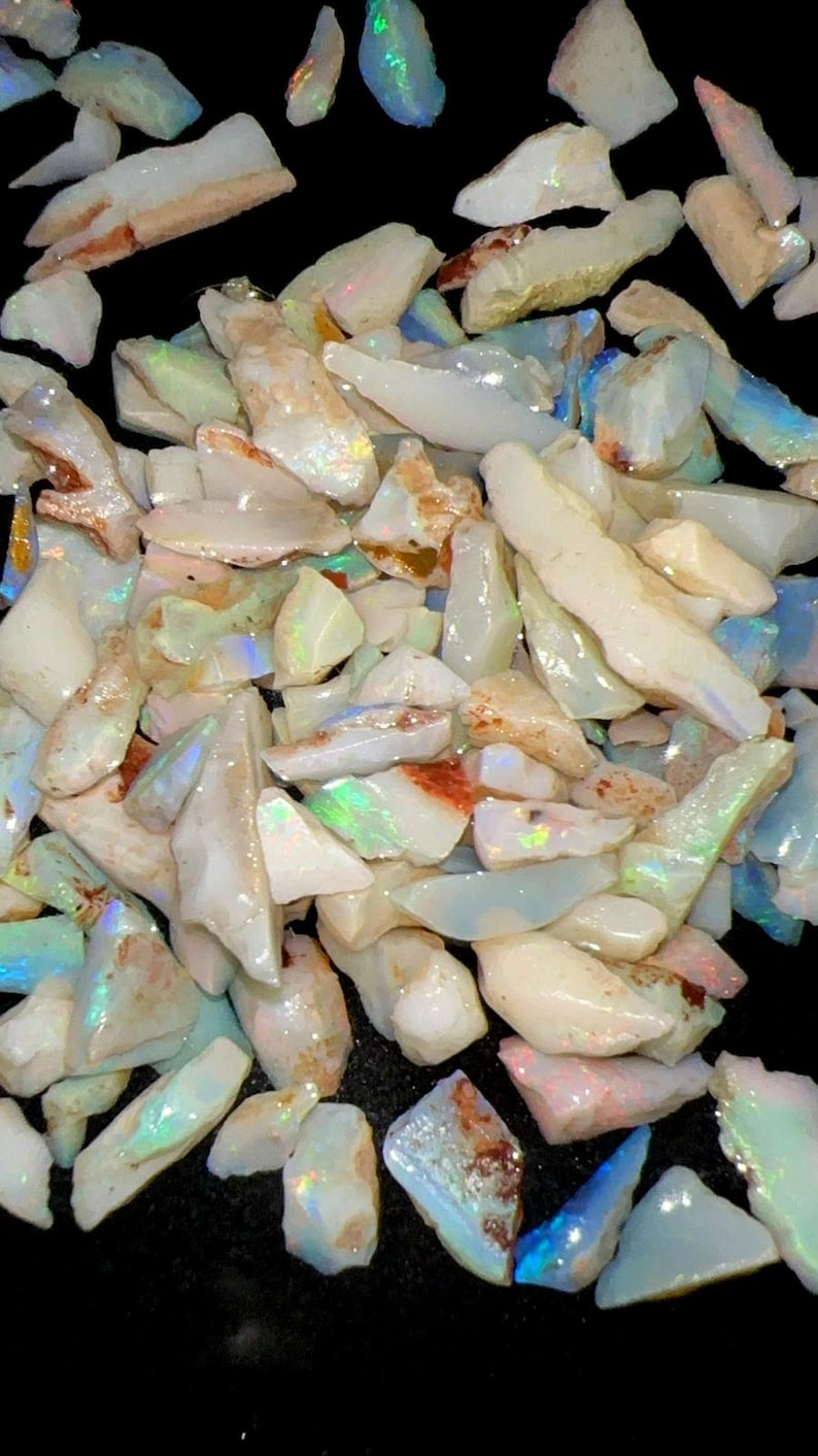 May include: A collection of raw, unpolished opal stones featuring a milky white base color with iridescent flashes of green, blue, and orange. The stones are irregular in shape and size, displaying a translucent to opaque texture with natural inclusions.
