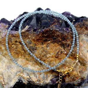 May include: A delicate necklace composed of small, faceted, translucent blue-grey beads arranged in a single strand. The necklace features a gold-toned metal lobster claw clasp and a short extender chain with small gold-toned beads at the ends.