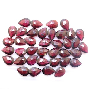 May include: A scattering of faceted, teardrop-shaped gemstones in shades of deep red. The gems are arranged closely together against a white background, highlighting their polished surfaces and internal facets. The overall effect is one of rich colour and sparkle.