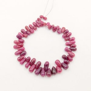 May include: A strand of polished, teardrop-shaped ruby beads. The beads are a deep, rich red color, with variations in tone. The beads are strung together on a thin, white string, and are arranged in a circular shape against a white background.