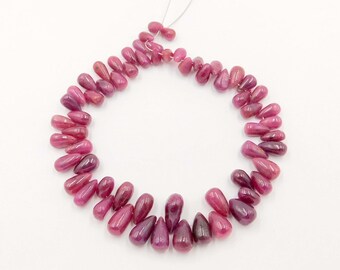 May include: A strand of polished, teardrop-shaped ruby beads. The beads are a deep, rich red colour, with variations in tone. The beads are strung together on a thin, white string, and are arranged in a circular shape against a white background.