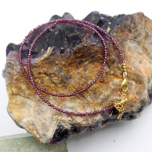 May include: A delicate necklace featuring tiny, faceted deep red garnet beads strung on a thin cord. The necklace includes a gold-toned metal lobster claw clasp and a short extender chain with small circular links for adjustable length.