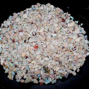 K&ouml;nnte beinhalten: Nahaufnahme eines Haufens kleiner, unregelm&auml;&szlig;ig geformter Opalchips. Die Chips sind &uuml;berwiegend cremefarben mit schillernden Farbt&ouml;nen, darunter Blau, Gr&uuml;n und Rot. Der Hintergrund ist schwarz.