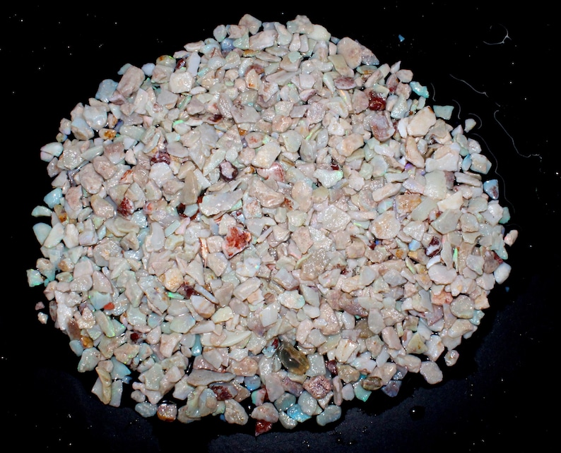 K&ouml;nnte beinhalten: Nahaufnahme eines Haufens kleiner, unregelm&auml;&szlig;ig geformter Opalchips. Die Chips sind &uuml;berwiegend cremefarben mit schillernden Farbt&ouml;nen, darunter Blau, Gr&uuml;n und Rot. Der Hintergrund ist schwarz.