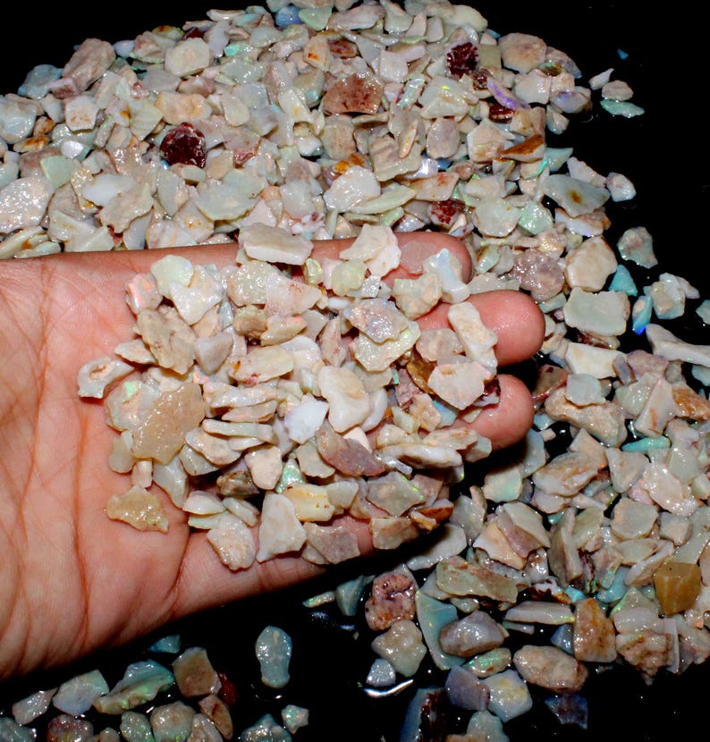 K&ouml;nnte beinhalten: Nahaufnahme einer Hand, die einen Haufen kleiner, unregelm&auml;&szlig;ig geformter Opalchips h&auml;lt. Die Chips zeigen eine Vielzahl von Farben, darunter Wei&szlig;, Beige und Hauch von Blau und Rot. Die Opale sind grob geschnitten und scheinen rohe Edelsteine zu sein.