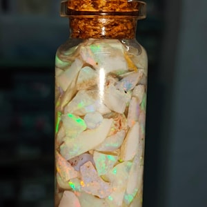 Könnte beinhalten: Eine transparente Glasflasche gefüllt mit schillernden Opalfragmenten. Die Flasche hat einen Korken. Die Opale zeigen eine Reihe von Farben, darunter Weiß, Grün und Orange. Die Flasche wird vor einem dunklen Hintergrund gehalten.