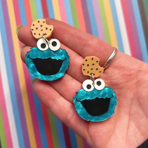 May include: A pair of novelty earrings featuring a blue monster face with a large black mouth and white eyes. Each earring has a small, tan cookie attached above the monster's head. The earrings are held in a hand against a colorful striped background.