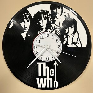 May include: A black vinyl record clock featuring a white silhouette of the band The Who. The clock face is white with black numbers and hands. The band's name is printed in white below the clock face.