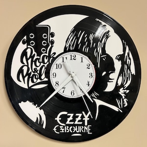 May include: Black vinyl record clock featuring a white silhouette of a guitar and the words "Rock & Roll". The clock face is white with black numbers and hands. The image also includes a black and white image of Ozzy Osbourne and his name.