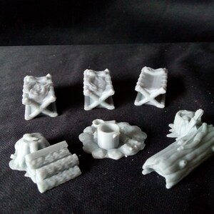 May include: A set of miniature camping accessories, including a campfire, a log, a bucket, and three wooden camp beds. The accessories are made of grey plastic and are designed for use in tabletop games or dioramas.