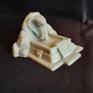 May include: A gray 3D printed miniature of a stone fountain with a cobra snake head pouring water into a basin. The fountain is on a square base with a number "8" on one side.